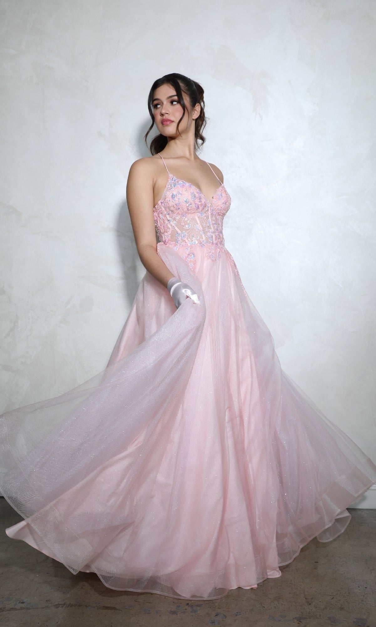A woman stands against a textured, light-colored wall wearing the Lace-Up Corset Long Prom Ball Gown: Eureka 9960, a light pink A-line dress with thin straps and floral embellishments.